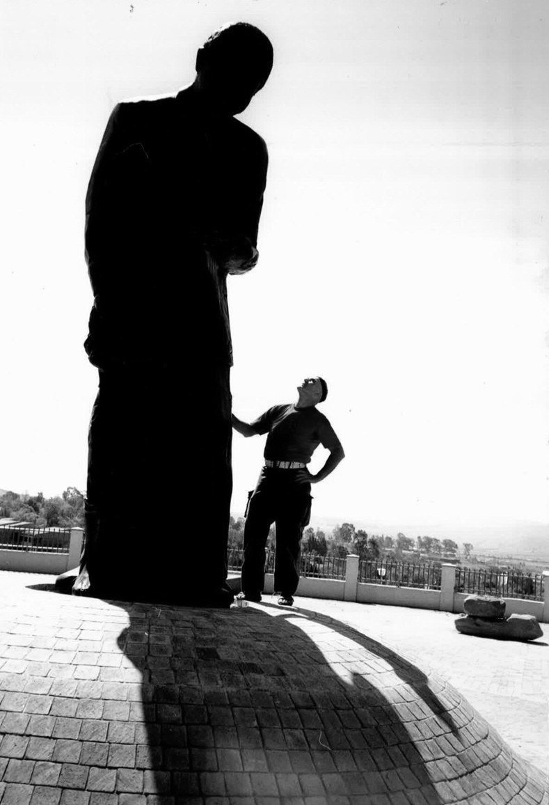 Kurt at the foot of the finished bronze, looking up — the silhouette of Madiba and Child with the Umtata sky behind it.