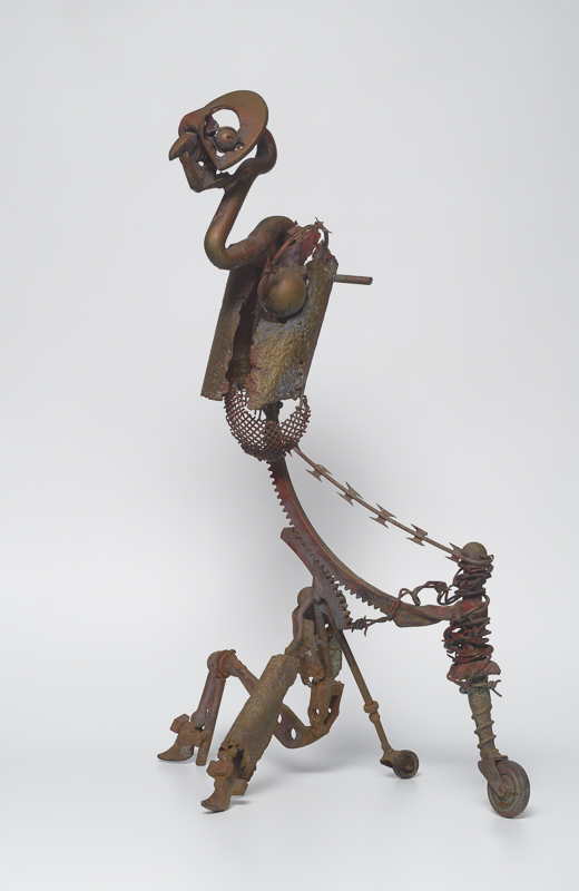 Lost Spirit — Welded scrap-metal figure in gold and red, beak-like mouth, large eyes, wheel-leg — gender in flux