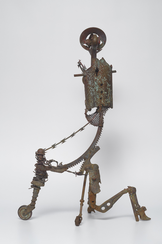 Lost Spirit — Welded scrap-metal figure in gold and red, beak-like mouth, large eyes, wheel-leg — gender in flux